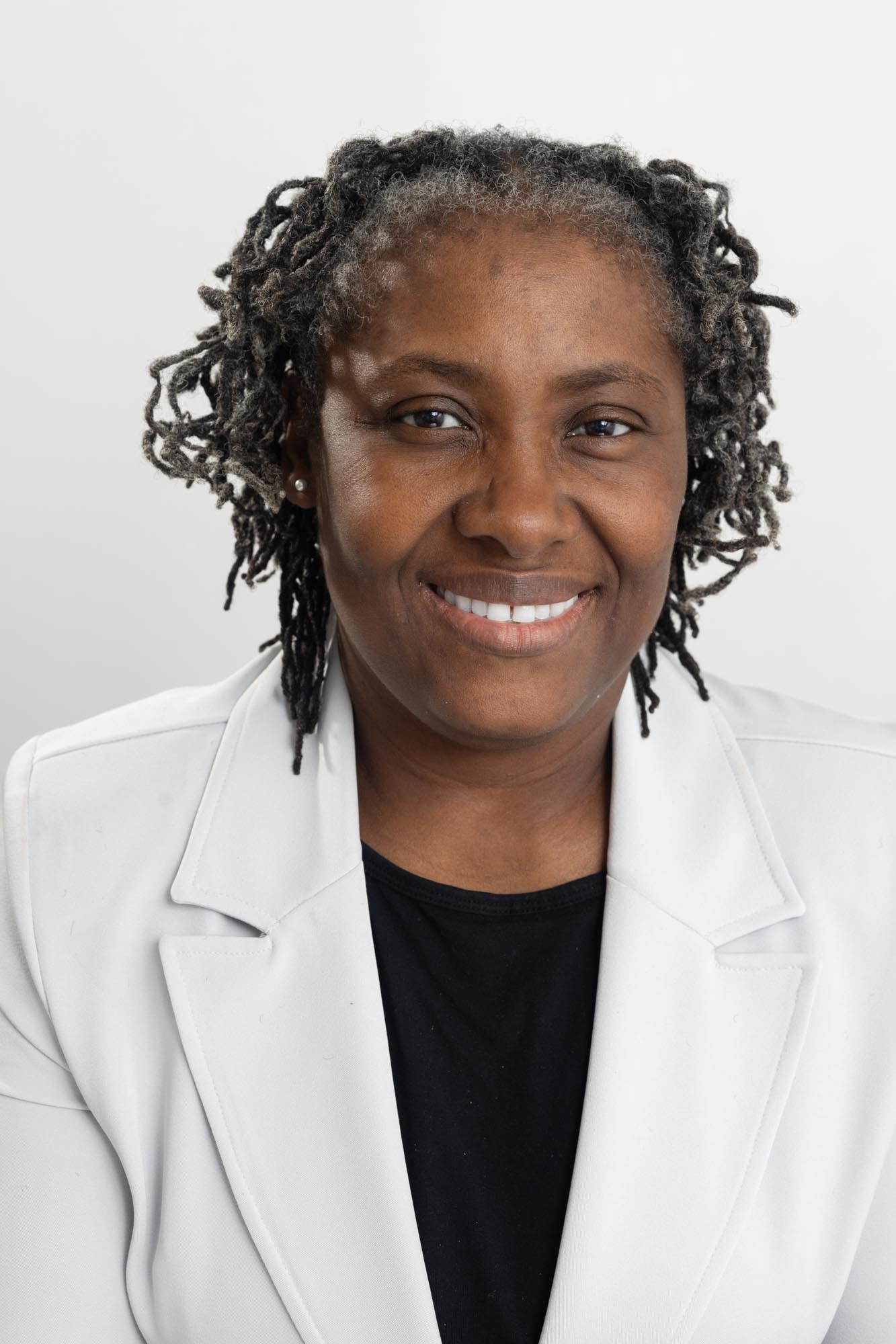 A Black woman with grey hair smiles. She is wearing a white jacket over a black blouse.